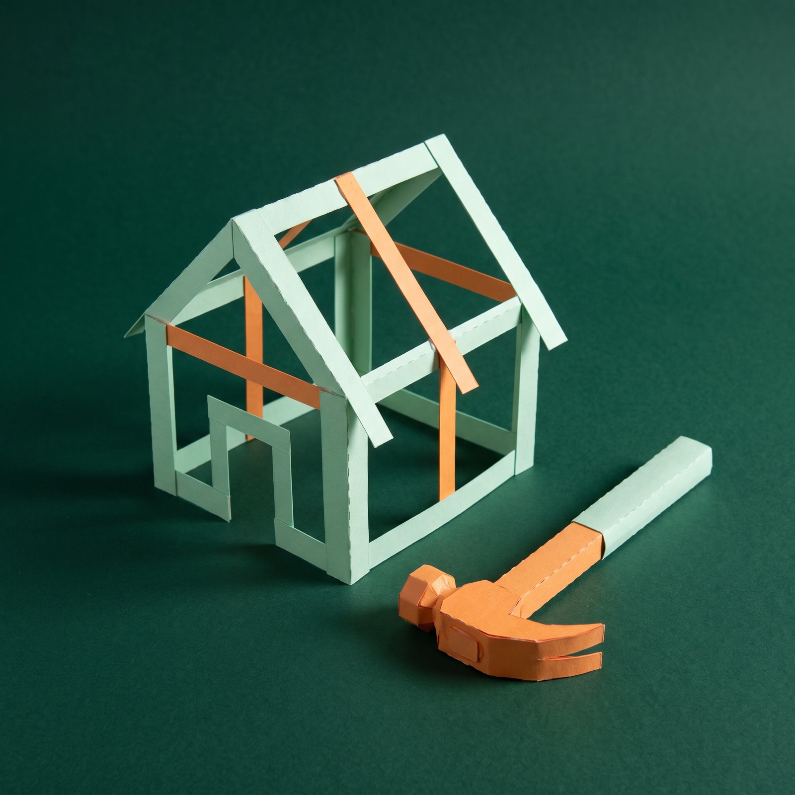 Paper sculpture of the timber frame of a small house under construction, with a claw hammer lying beside it