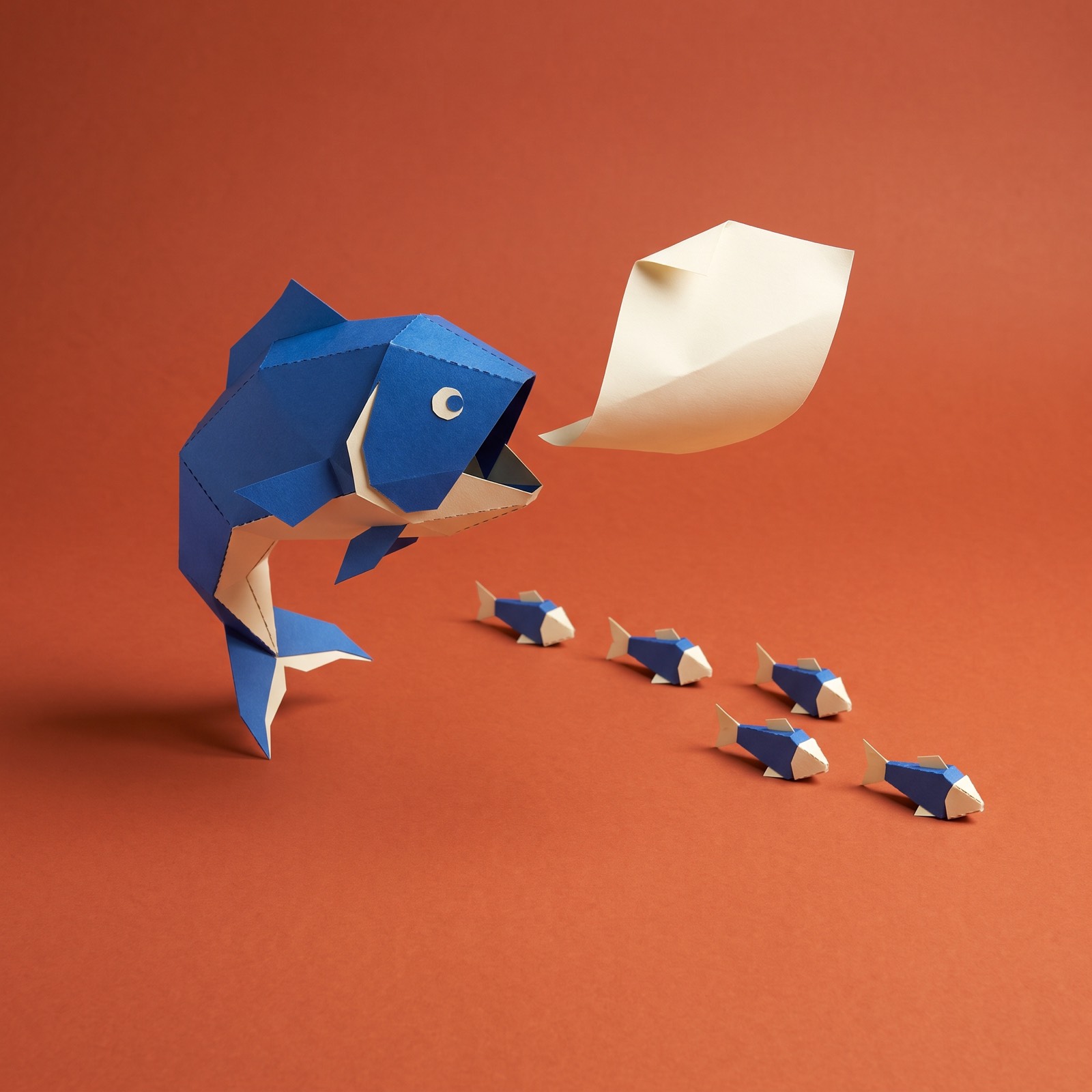 Paper sculpture of a very large fish chasing a line of small fish