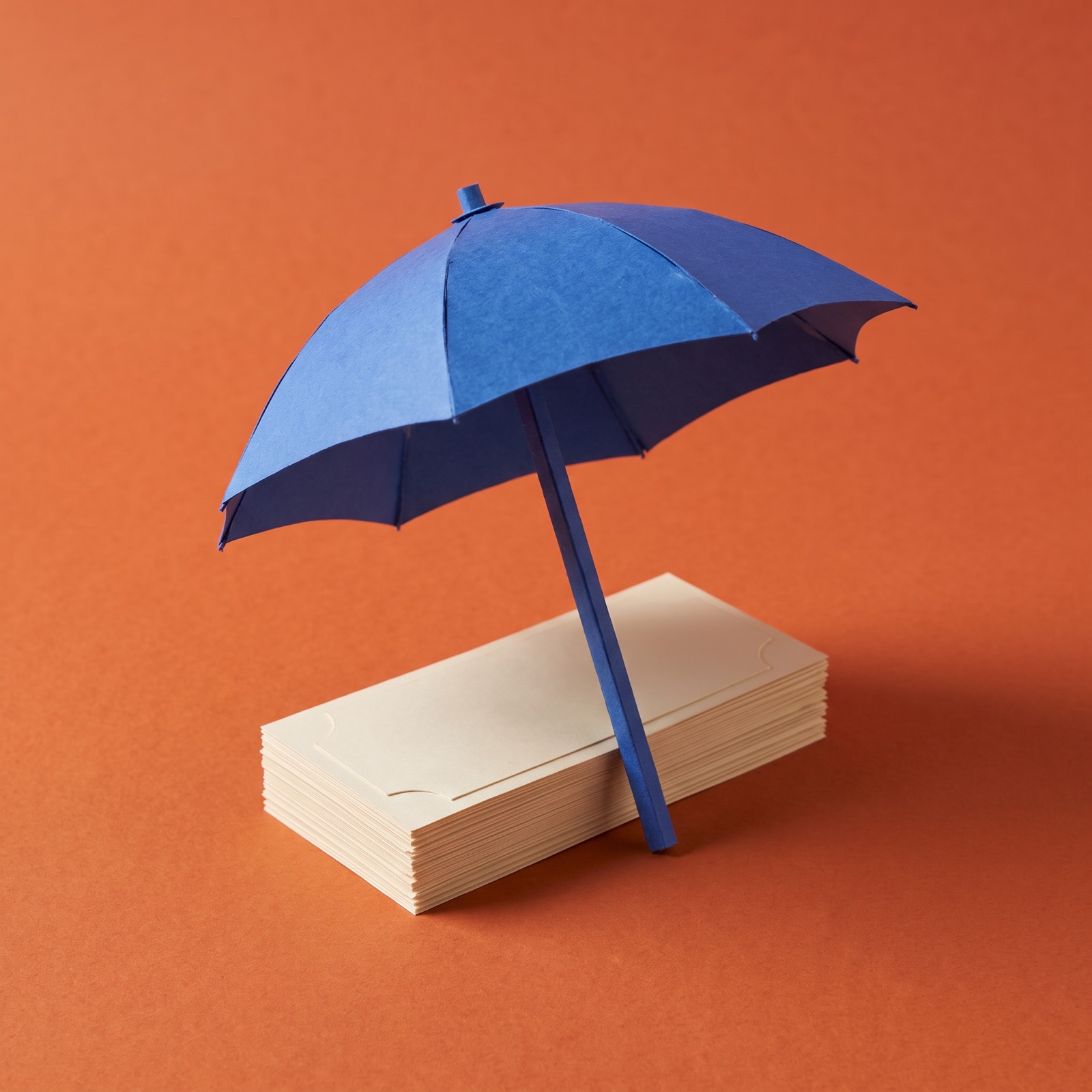 Paper sculpture of a small open umbrella standing over a neat stack of banknotes, sheltering it